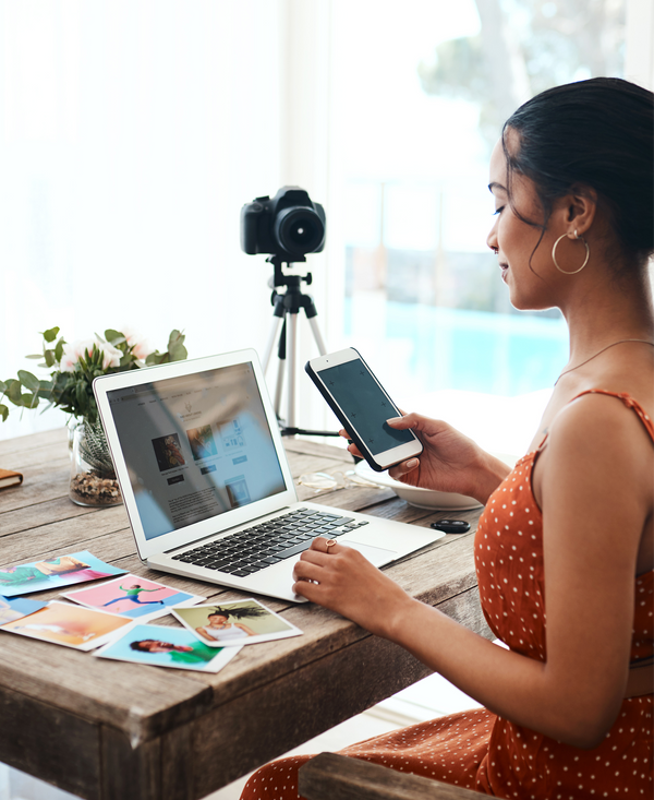 A creative workspace with a MacBook showing photo editing grid, surrounded by cameras, magazines, and a person taking a photo with a phone.